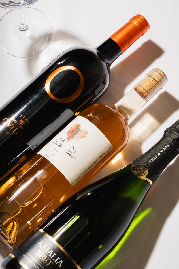 Three bottles of Greek wine laid on a white table beside a wine glass (official image)