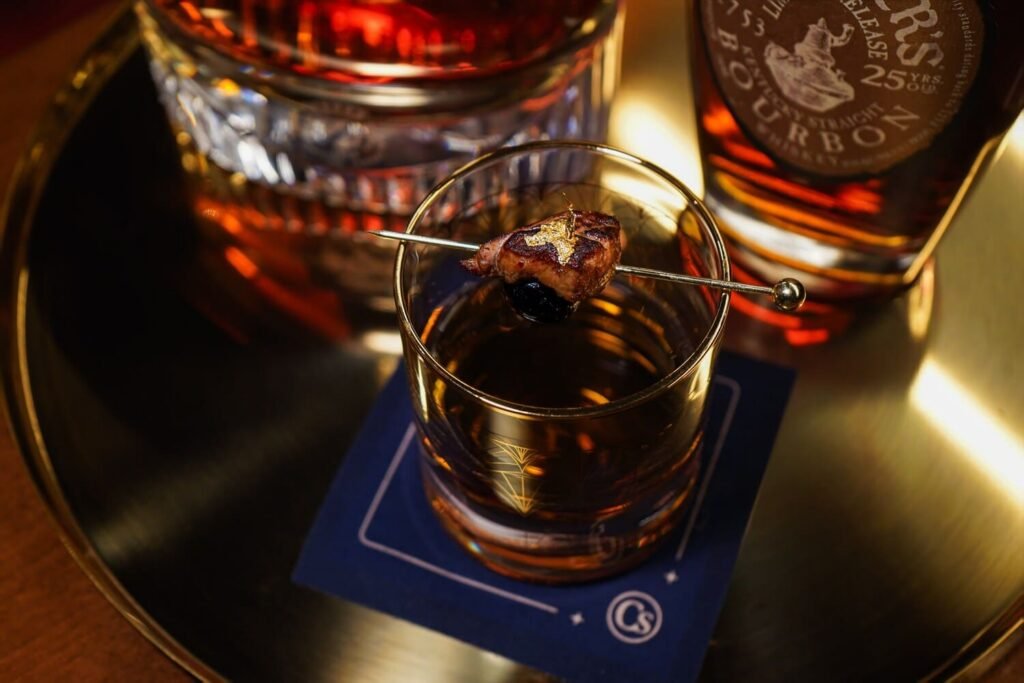 A bourbon cocktail on a navy coaster beside a bottle of Michter’s at Carversteak (official photo)