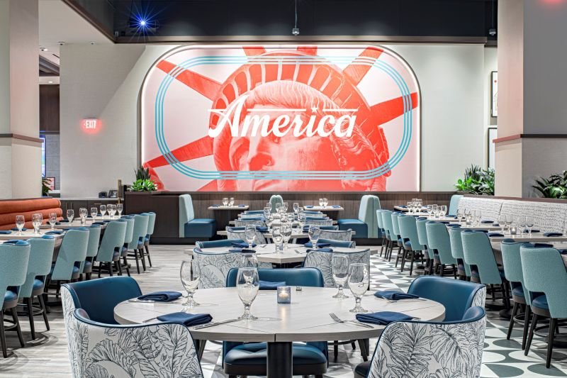 Dining room at America with blue chairs, set tables and a big red Statue of Liberty mural with the word America (venue photo by Chris Wessling)