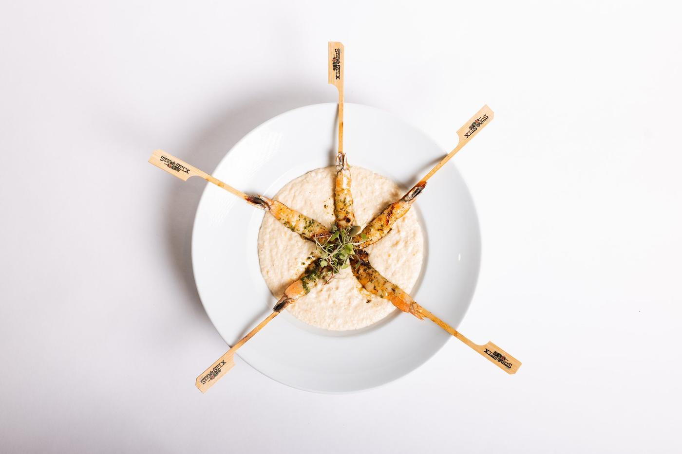Five grilled shrimp on branded Stew’s Stix paddles arranged over creamy grits in a white bowl with microgreens (venue photo)