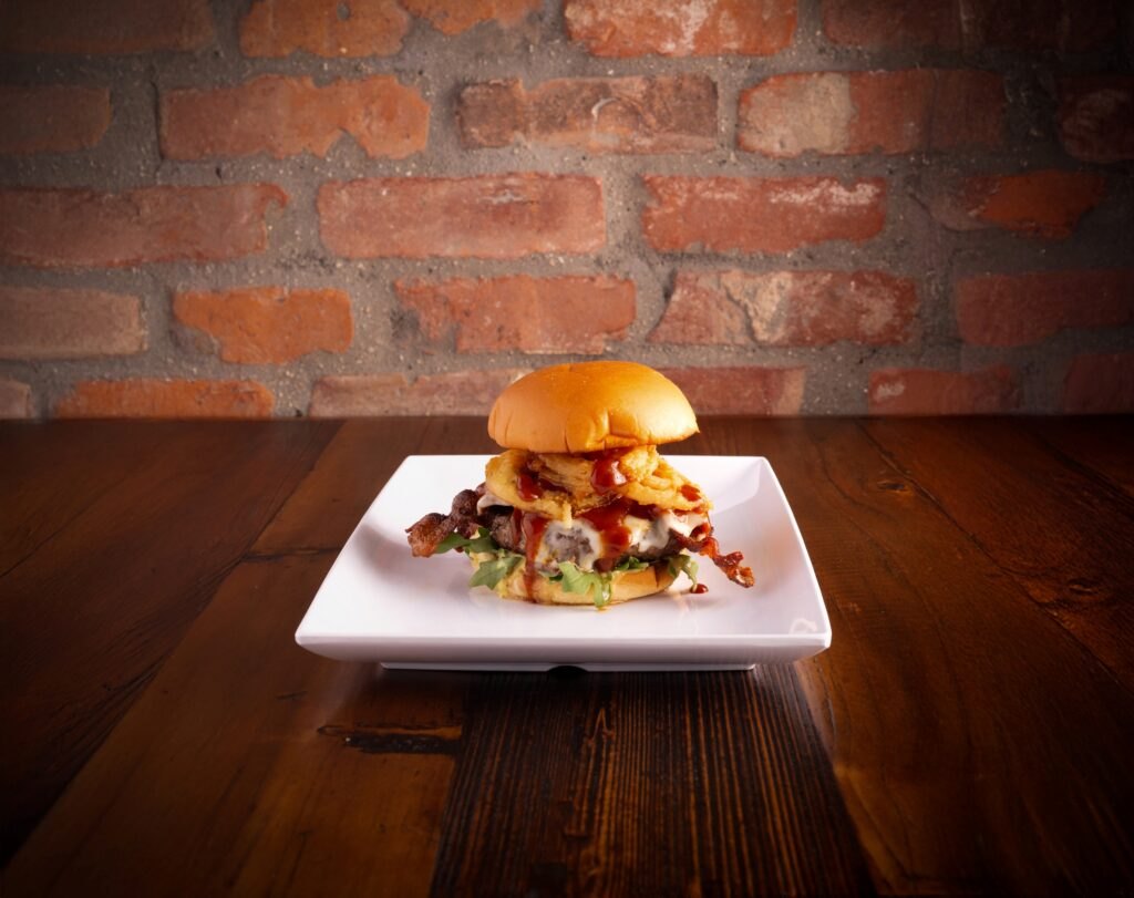 Burger topped with bacon and onion rings on a white plate in front of a red brick wall (Finney's Crafthouse photo)