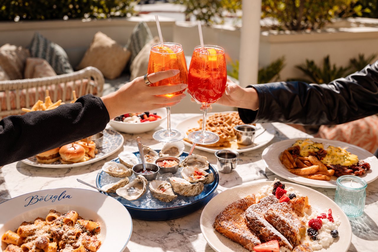 Two hands clinking orange spritzes over a patio brunch table with oysters, French toast, rigatoni and waffles (Bottiglia photo)