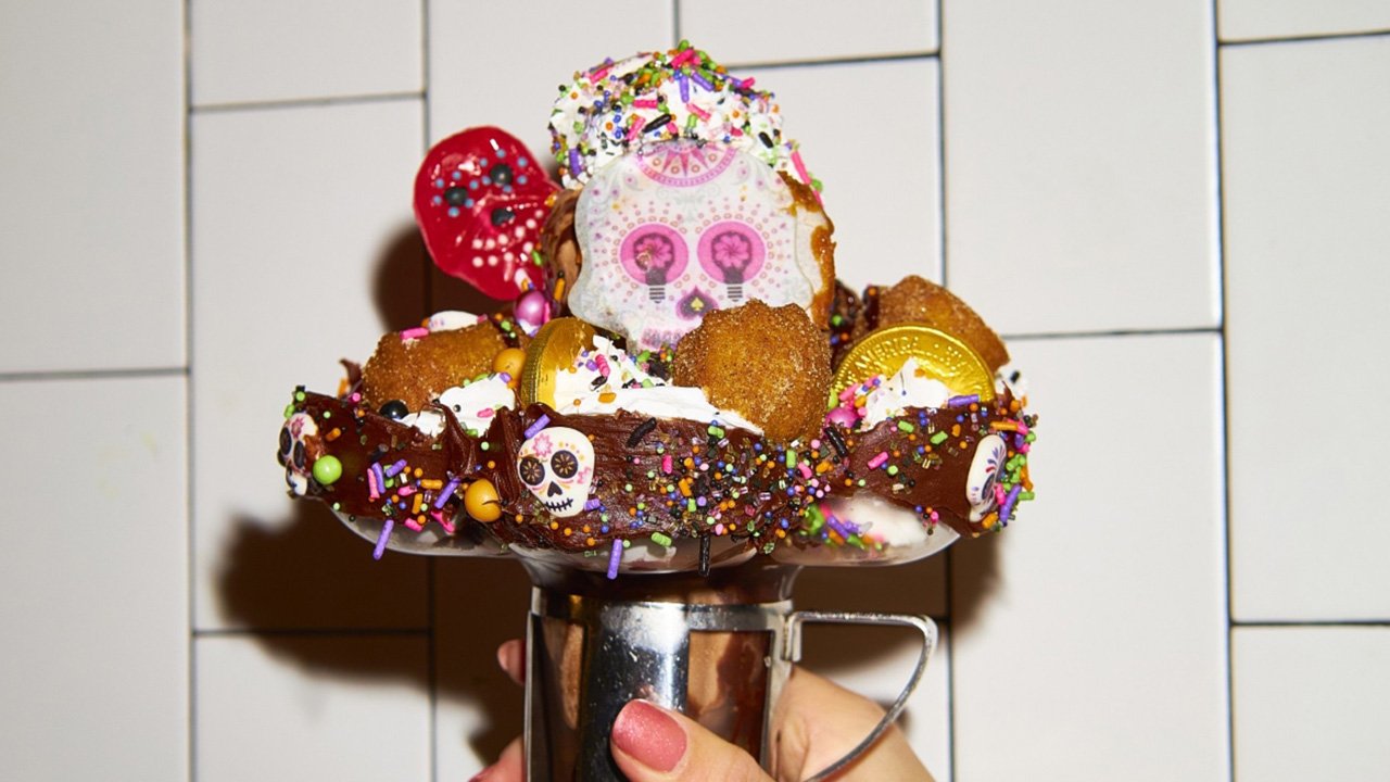 A hand holding up Black Tap's Día de los Muertos sundae, piled with churro bites, gold coins and a sugar-skull cookie (Black Tap photo)
