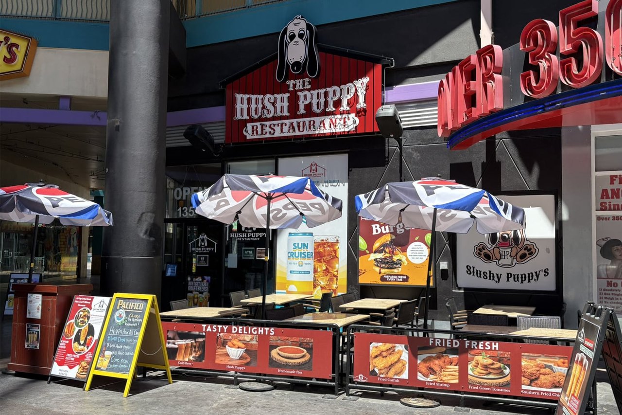 The Hush Puppy Restaurant storefront at Neonopolis with a hound-dog sign, patio umbrellas and fried-food menu banners (Vital Vegas photo)