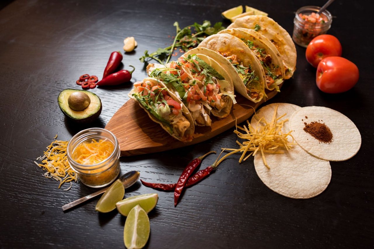 A row of tacos on a wooden board with lime wedges, dried chiles, tomatoes and tortillas (Borracha photo)